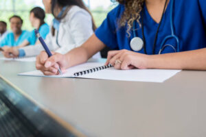 An unrecognizable medical student sits at a table in her lecture hall and writes in her class workbook.