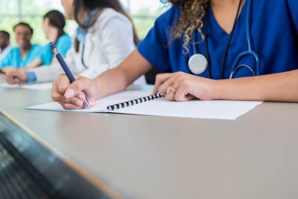 An unrecognizable medical student sits at a table in her lecture hall and writes in her class workbook.